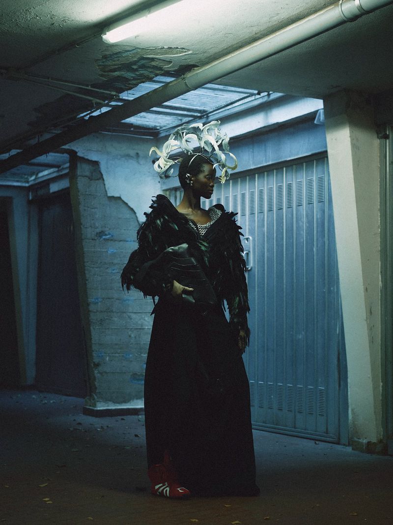 Woman in dark gown with feather epaulettes and headpiece, standing in garage, lit by fluorescent light. Fashion editorial photography.