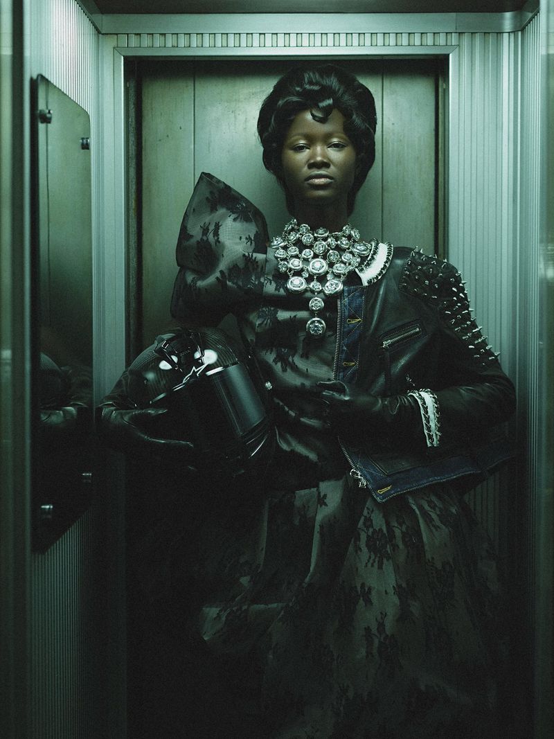 An adult Black woman is shown from the chest up in an elevator. She wears a jewel-covered dress, leather jacket, and gloves. Editorial shot.
