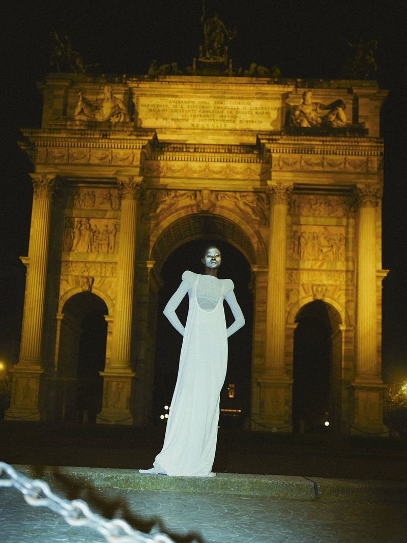 Woman in a long white dress posing in front of the Arco della Pace at night, illuminated by spotlights.