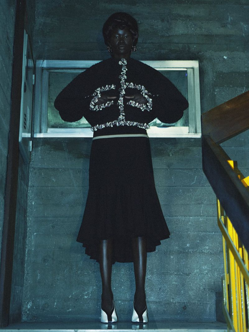 An African-American woman models a black skirt and sweater while standing on a concrete stairwell with a yellow railing, in a studio.