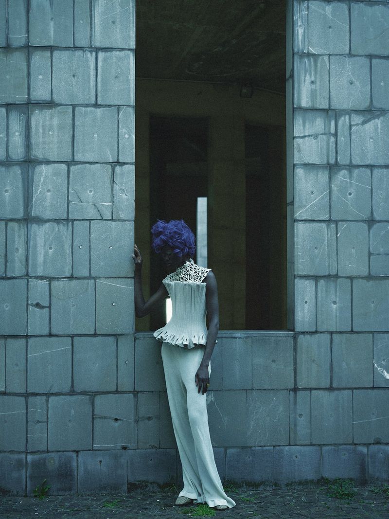 Fashionable person in pale suit posing in a derelict space with cinder block walls, wearing a detailed lacy bodice and bright violet wig.