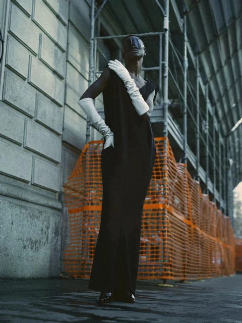 Elegant woman in a black dress and long white gloves posing in an urban setting with scaffolding in the background.