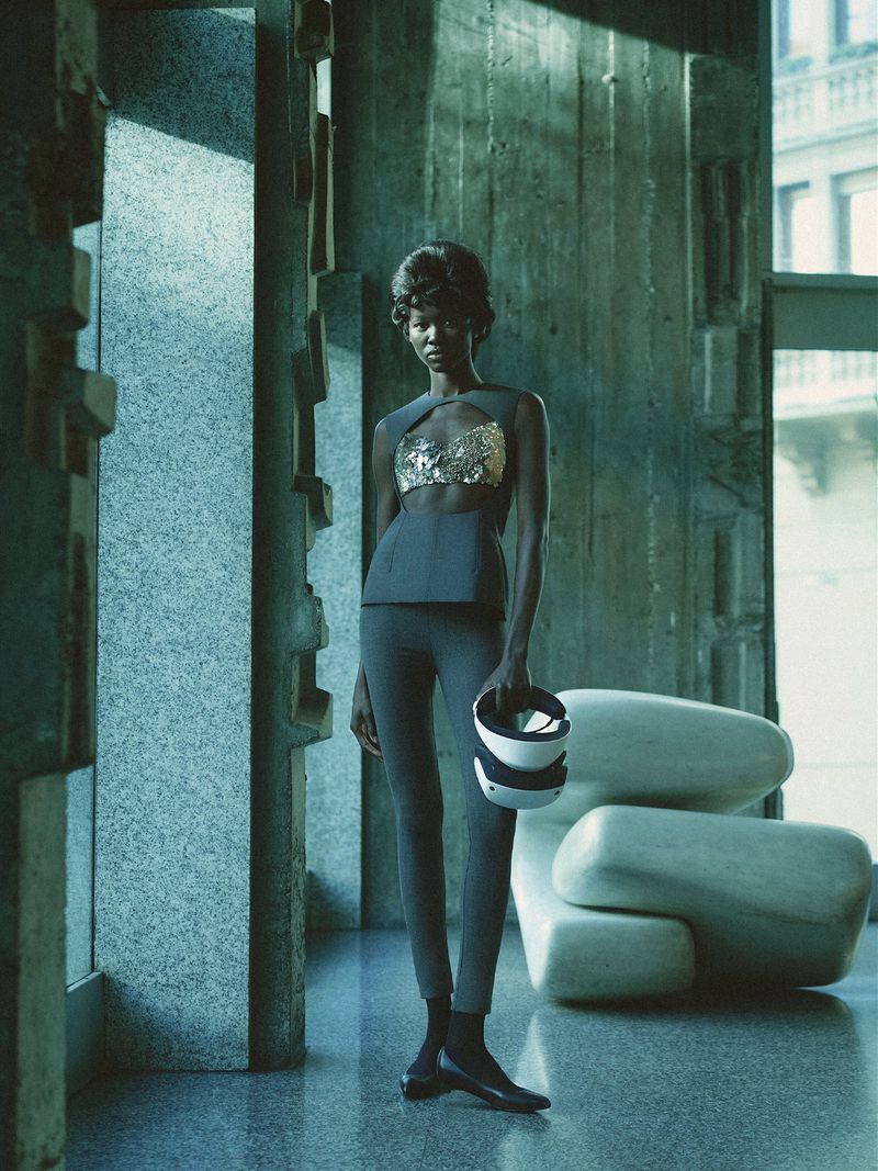 Woman in modern fashion stands in a textured concrete building interior with natural light. She holds VR headset with sculptural chair.