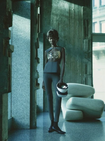 Woman in modern fashion stands in a textured concrete building interior with natural light. She holds VR headset with sculptural chair.