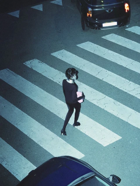 Woman in black clothes crossing crosswalk at night, cars nearby. High angle view of pedestrian safety and urban scene.