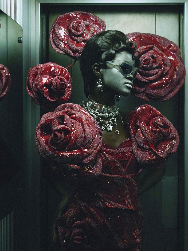 Elegant black woman poses in metallic elevator with dramatic rose-embellished dress and jewelry, wearing retro sunglasses for fashion editorial.