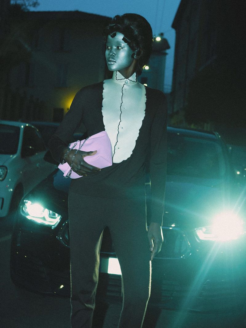 Fashionable woman with metallic makeup poses at night in front of parked cars. She wears a dark sweater, pants, and a light pink clutch.