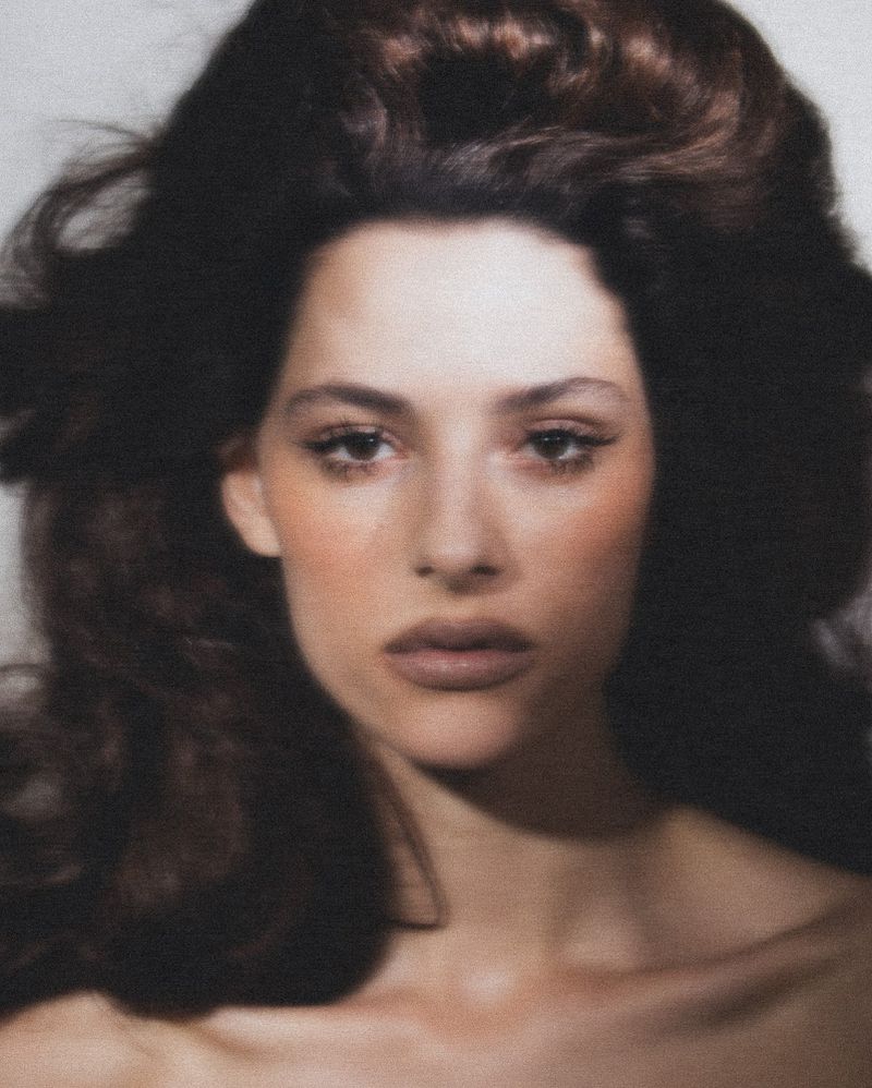 Close-up portrait of a woman with brown hair styled in large waves, wearing minimal makeup, against a plain white background.