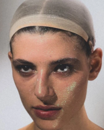 Close-up of a woman with dark eyeshadow and a tan wig cap, showcasing a beauty or fashion aesthetic against a neutral backdrop.