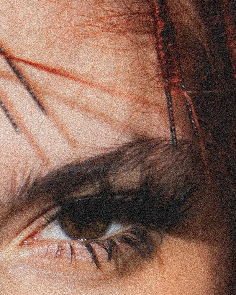 Extreme close-up of a woman's eye with brown iris, dark eyebrow, fair skin, and eyelashes with hints of makeup. Grainy texture.