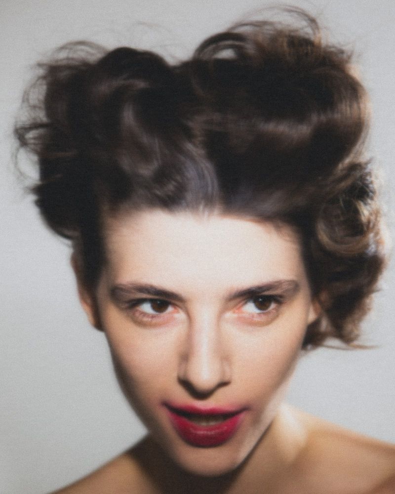 Close-up portrait of a fair-skinned adult female with dark, styled hair and red lipstick, set against a neutral, light-toned background.