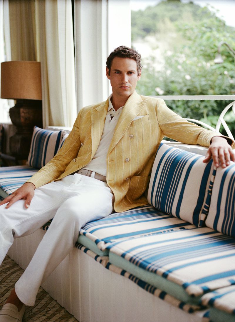 A casually-posed man in a yellow pinstripe blazer and white pants sits on a striped blue and white sofa in a well-lit indoor setting.