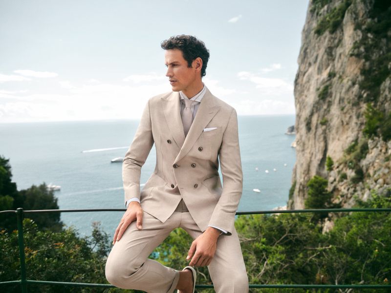 Man in a beige double-breasted suit sits on a metal railing with a coastal view featuring cliffs and the ocean in the background.