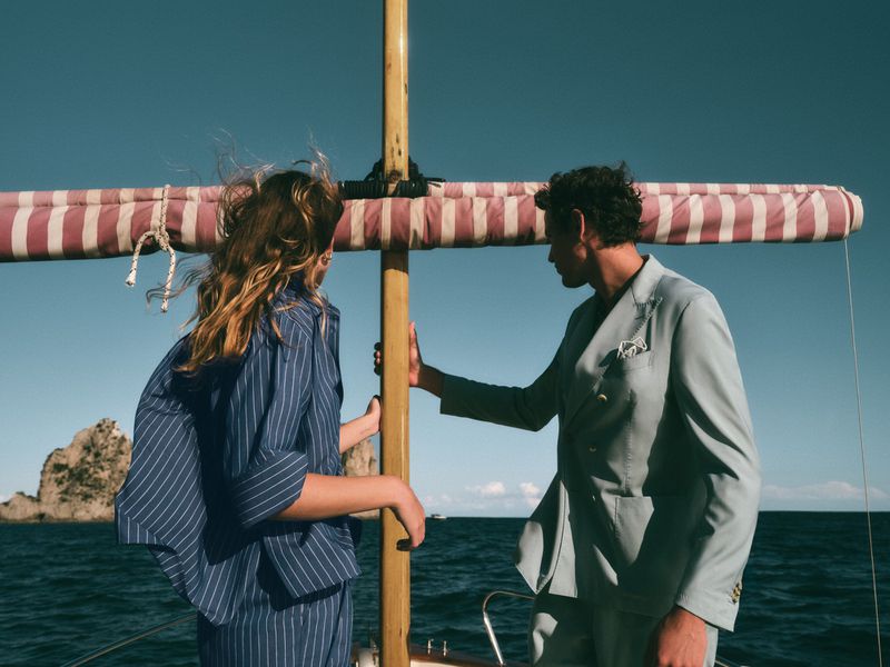A woman and a man in stylish suits stand on a boat with a striped awning, with the sea and a rocky island in the background.