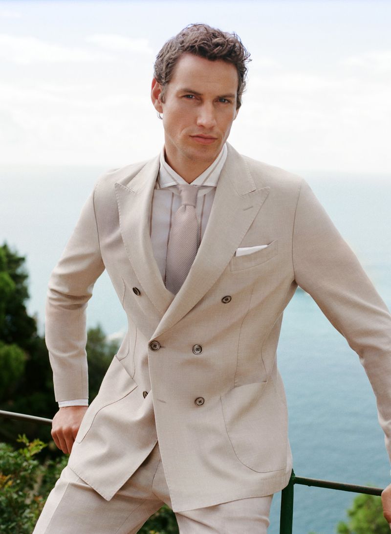 Man in a beige double-breasted suit leans on a railing with a distant ocean view, exuding sophisticated style and timeless elegance.