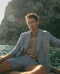 A man in a light blue suit and striped polo sitting on a boat with rocky cliffs and ocean in the background.