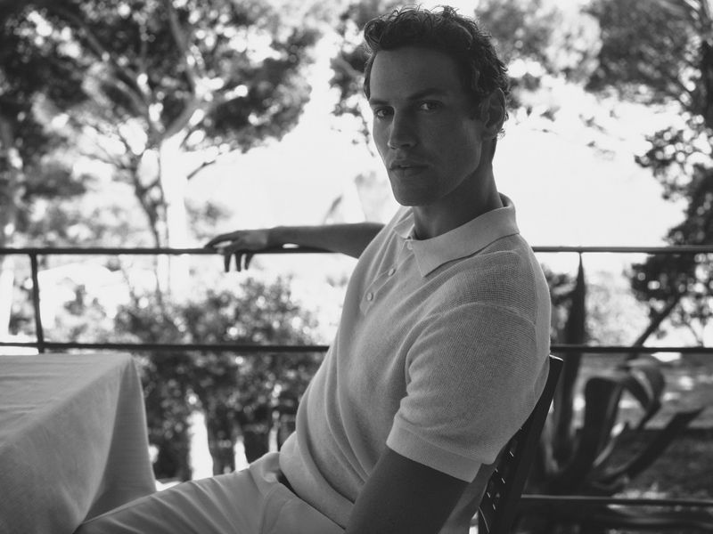 Monochrome portrait of a male model in a polo shirt seated outdoors against a railing and trees for Windsor spring summer 2025 campaign