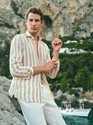 Male model wearing a striped shirt and white trousers leaning on rocks by the water for Windsor