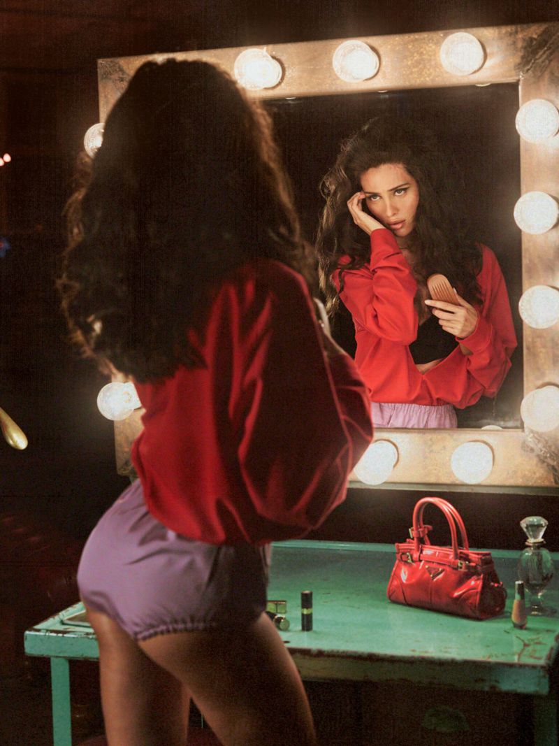 Woman in red sweater and lilac shorts looks in a vanity mirror at her reflection in an indoor setting with makeup on a table.
