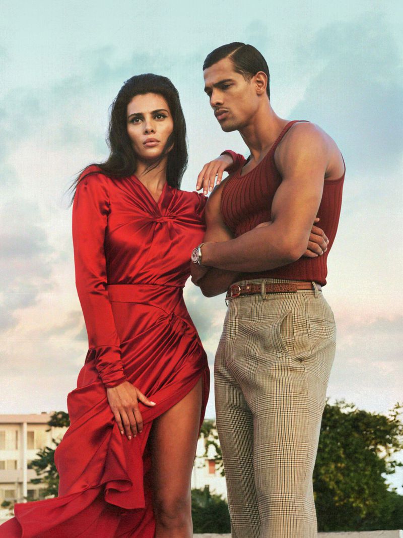 Attractive couple poses outdoors near building. Woman wears a long red dress with a high slit; man wears a maroon tank top and plaid pants.