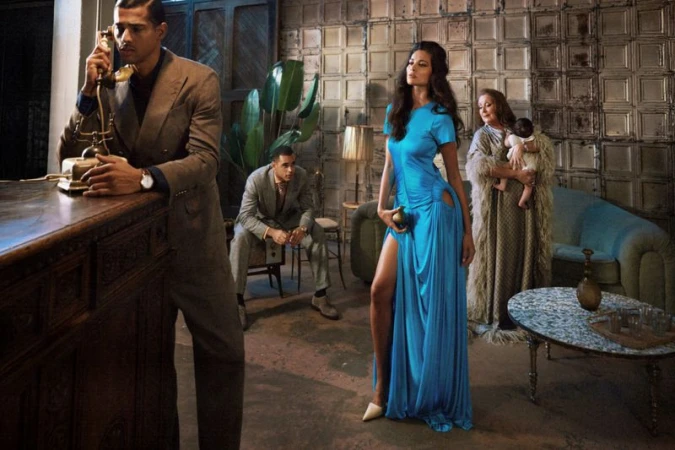 Fashion editorial shot indoors featuring an adult woman in a blue dress, plus three other people and vintage furniture in the background.