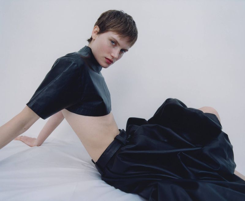 Portrait of a young adult female with a pixie haircut wearing a black crop top and draped skirt, posing against a white backdrop.