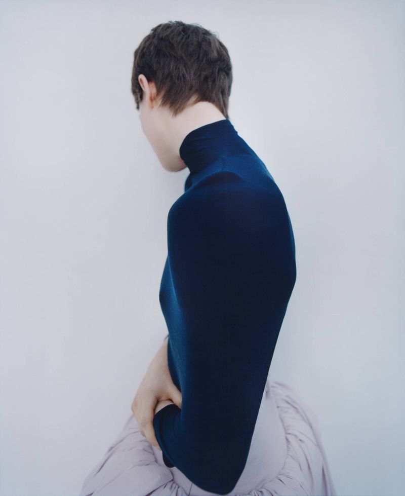 Back view of adult with short brown hair wearing blue turtleneck sweater and draped skirt against off-white background in portrait orientation.