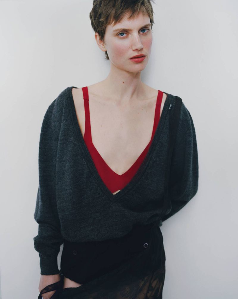Young woman poses indoors wearing gray sweater, red top, and black skirt against a white wall in a fashion editorial shot.