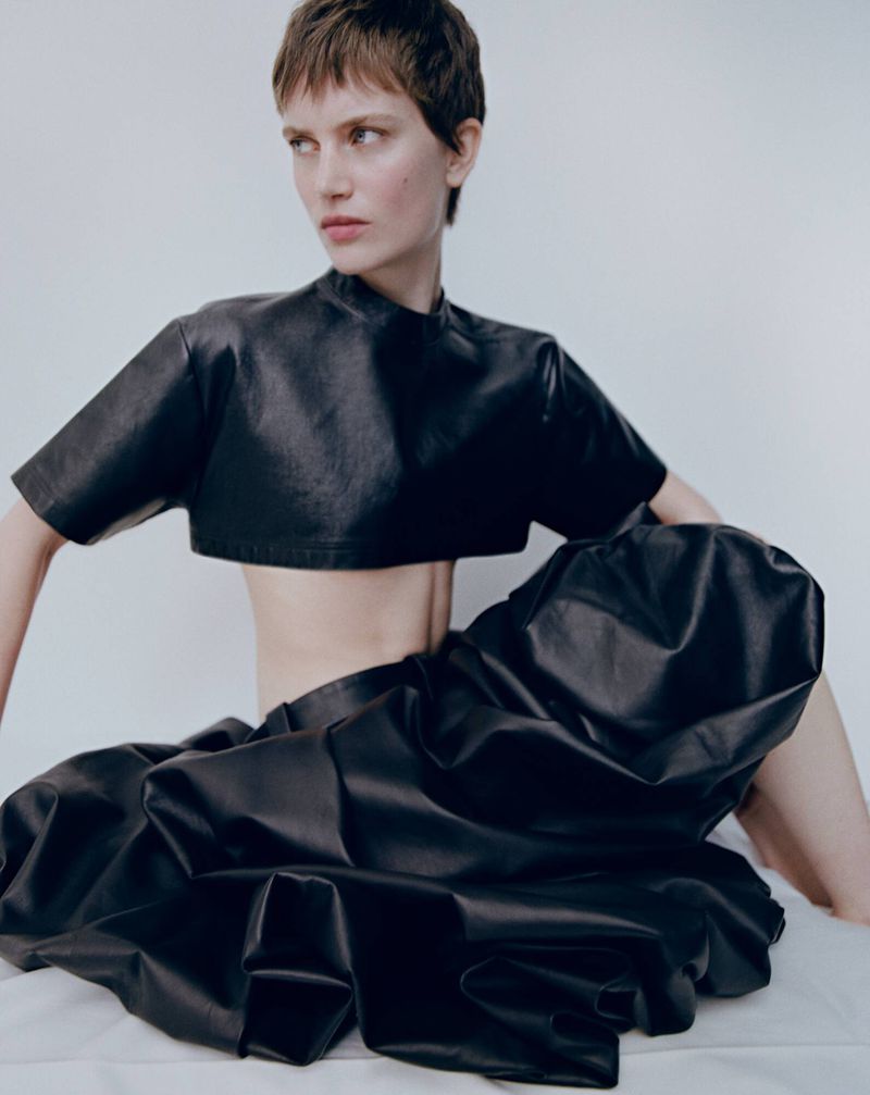 Woman poses in a studio wearing a black leather crop top and a voluminous skirt. The studio backdrop is white, and the lighting is bright.
