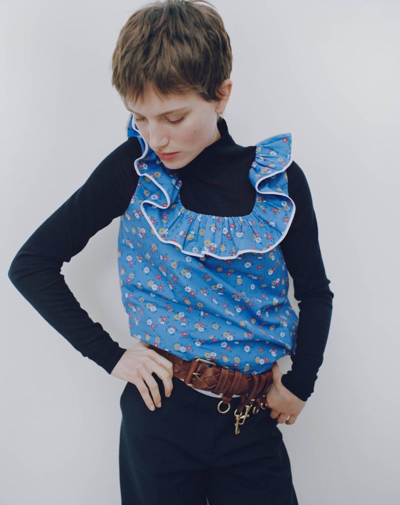 A young adult woman poses in a studio wearing a blue floral top with ruffles, black long-sleeve turtleneck, black pants and a brown belt.