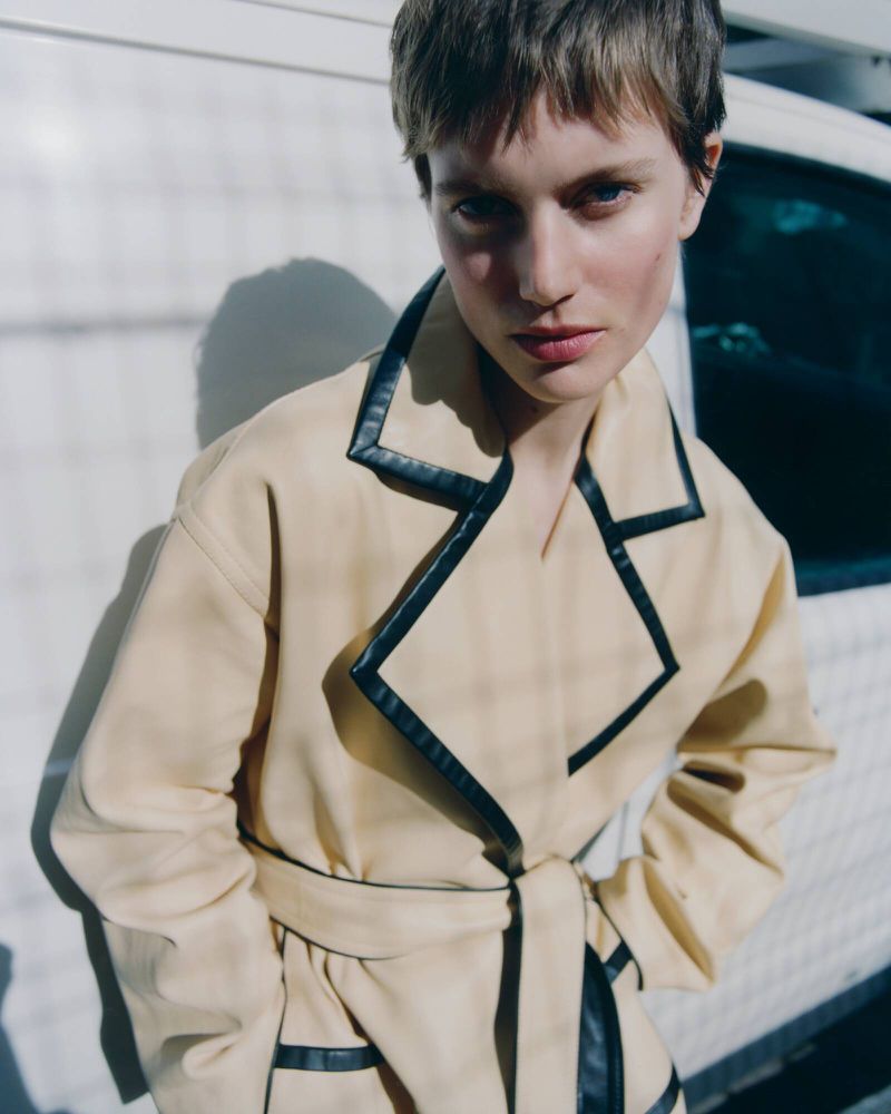 Close-up of a woman with short brown hair, wearing a beige trench coat with black trim, posing outdoors in natural sunlight.