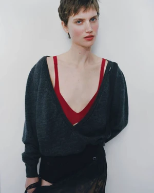 Model with short brown hair wearing a dark grey V-neck sweater over a red top against a white seamless background.