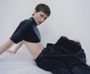 Model with short brown hair wearing a black leather crop top and dark trousers seated against a white background
