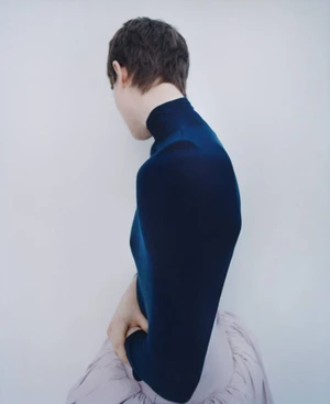 Model with short brown hair wearing a dark blue turtleneck and pale pink skirt against a white wall.
