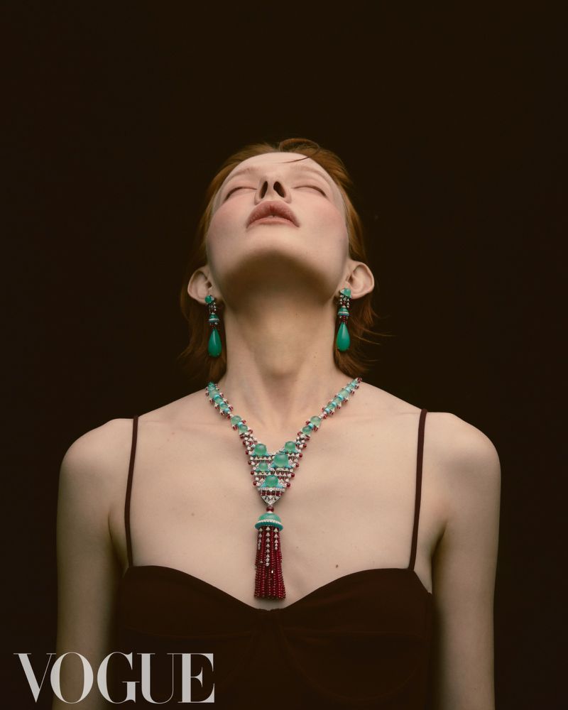 Fashion shot of a redhead woman with eyes closed wearing an elaborate turquoise and ruby necklace and matching earrings against a dark backdrop.