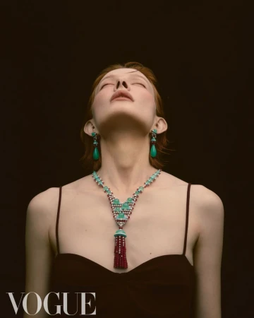 Fashion shot of a redhead woman with eyes closed wearing an elaborate turquoise and ruby necklace and matching earrings against a dark backdrop.