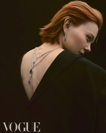 Elegant fashion shot of a red-haired woman wearing a black dress and sapphire jewelry against a dark background. Vogue logo visible.