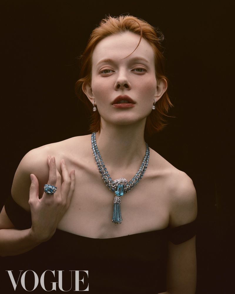 Portrait of red-haired woman wearing an off-the-shoulder black dress, necklace, ring, and earrings against a dark background. Vogue logo visible.