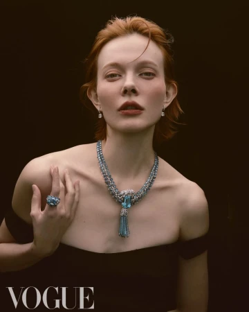 Portrait of red-haired woman wearing an off-the-shoulder black dress, necklace, ring, and earrings against a dark background. Vogue logo visible.