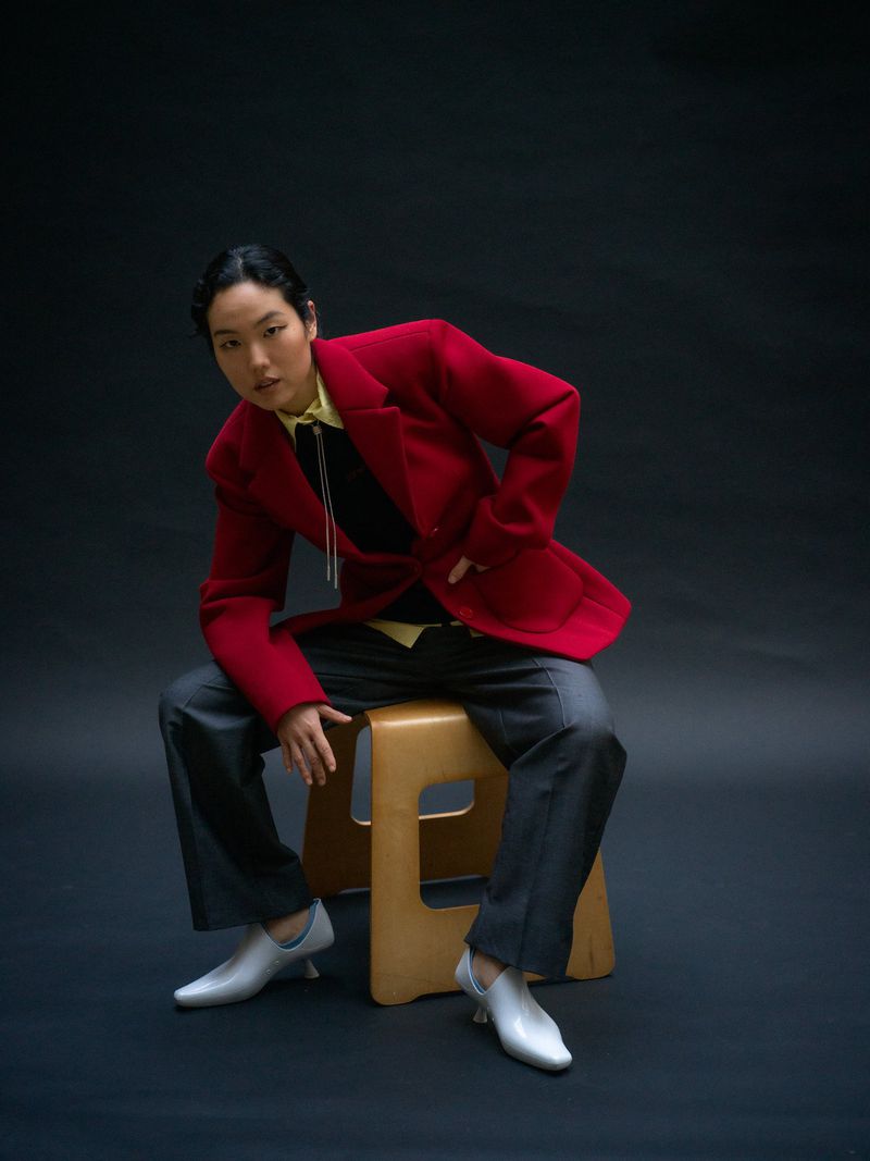Stylish person in red jacket poses seated on a stool in a studio with dark background. The focus is on their outfit and confident pose.