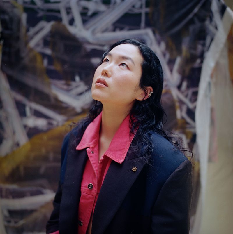 Portrait of an Asian adult female looking up, wearing a pink collared shirt and a blue blazer against a painted abstract background.