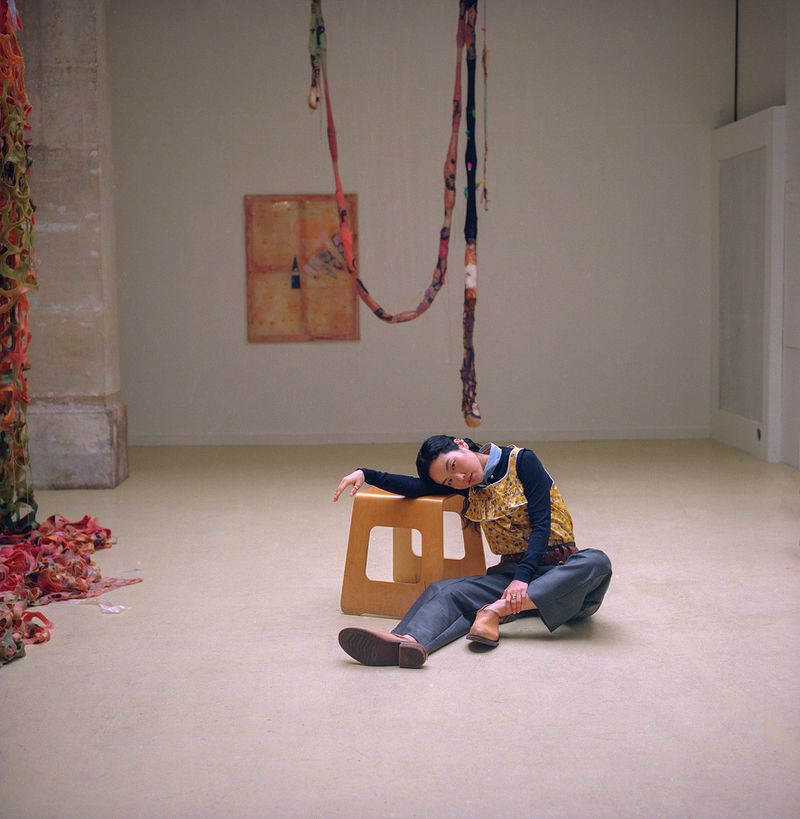 Woman rests on a stool in an art gallery surrounded by installation art, wearing a mustard-yellow top and grey pants in a spacious room.