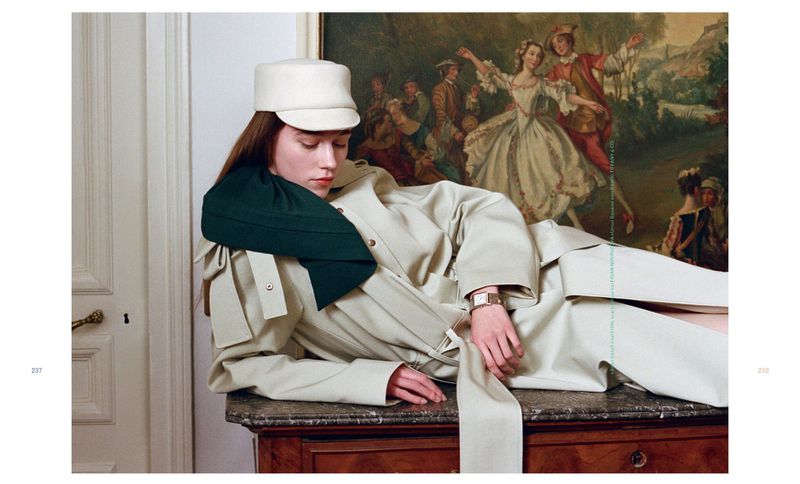 Fashion editorial shot of a woman in a trench coat and hat, lying on a wooden dresser in front of a classical painting, indoors.