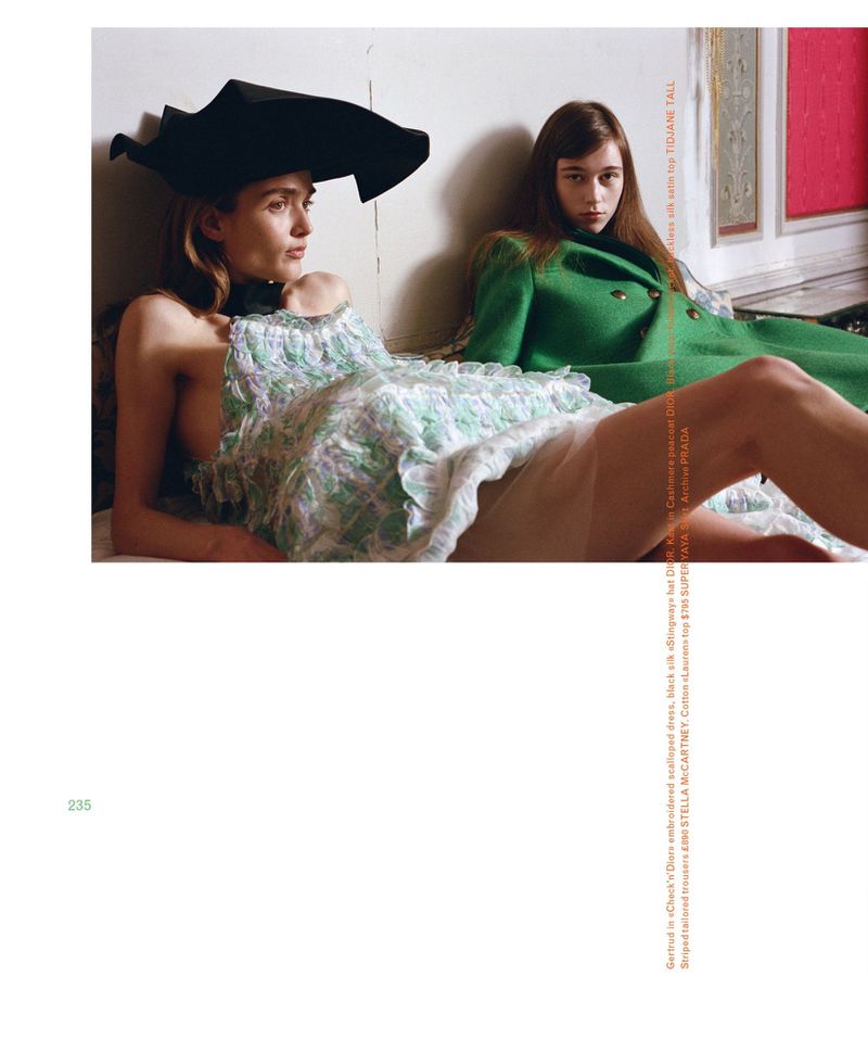 Two women in a fashion editorial. The first woman wears a large black hat and a light dress. The second wears a green coat.