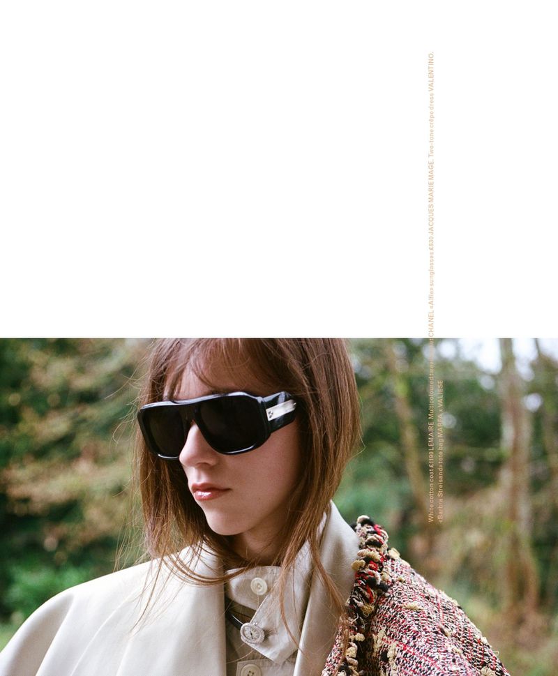A woman with bobbed brown hair wearing black sunglasses, a beige coat, and a tweed jacket stands against an outdoor green backdrop.