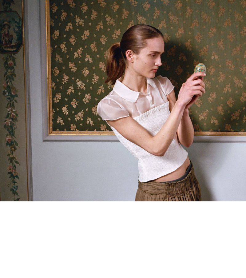 Woman in white top holds decorated egg indoors with floral wallpaper backdrop, bathed in diffused natural light in a neutral setting.