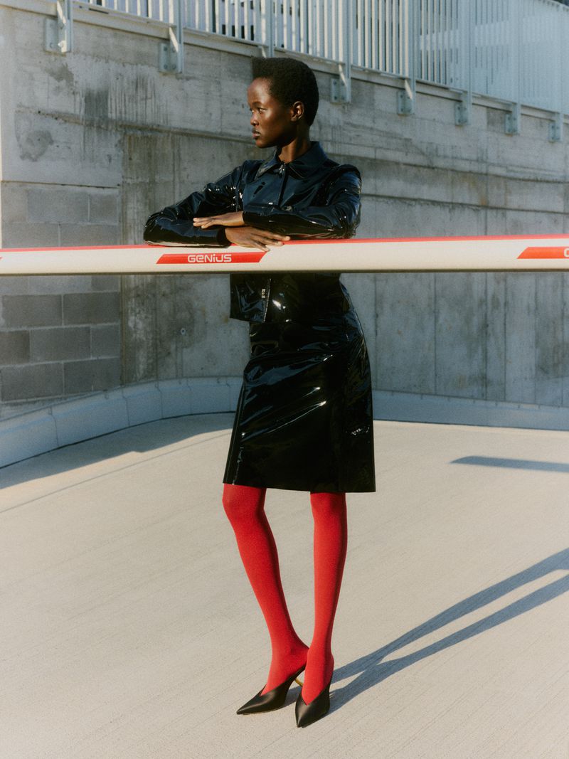 Elegant Black woman posing in glossy jacket and skirt with red tights in an urban setting, showcasing fashion and style with modern flair.
