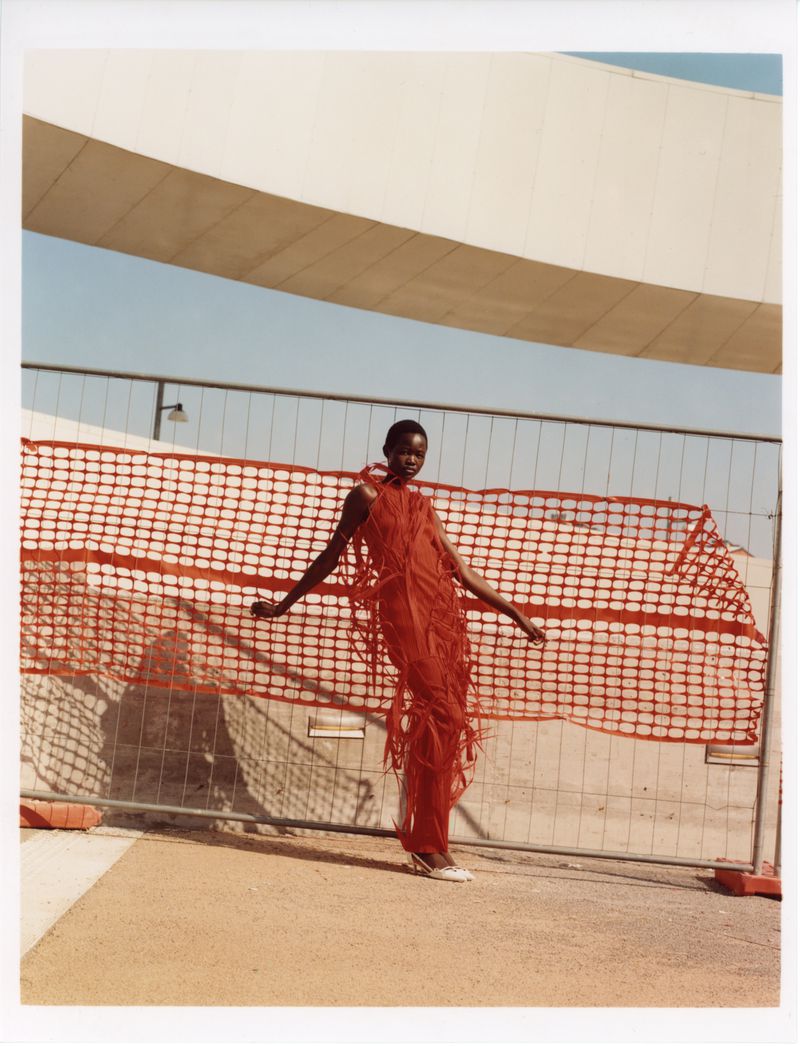 A tall, dark-skinned woman poses in an airy red dress with fringe, set against a backdrop of a modern bridge and fence.