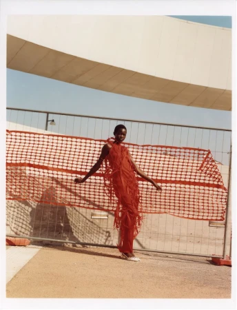 A tall, dark-skinned woman poses in an airy red dress with fringe, set against a backdrop of a modern bridge and fence.