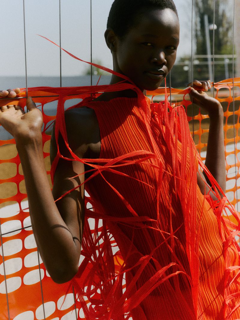 Adult female fashion model poses in an orange dress with fringe against an orange safety fence outdoors during daytime.
